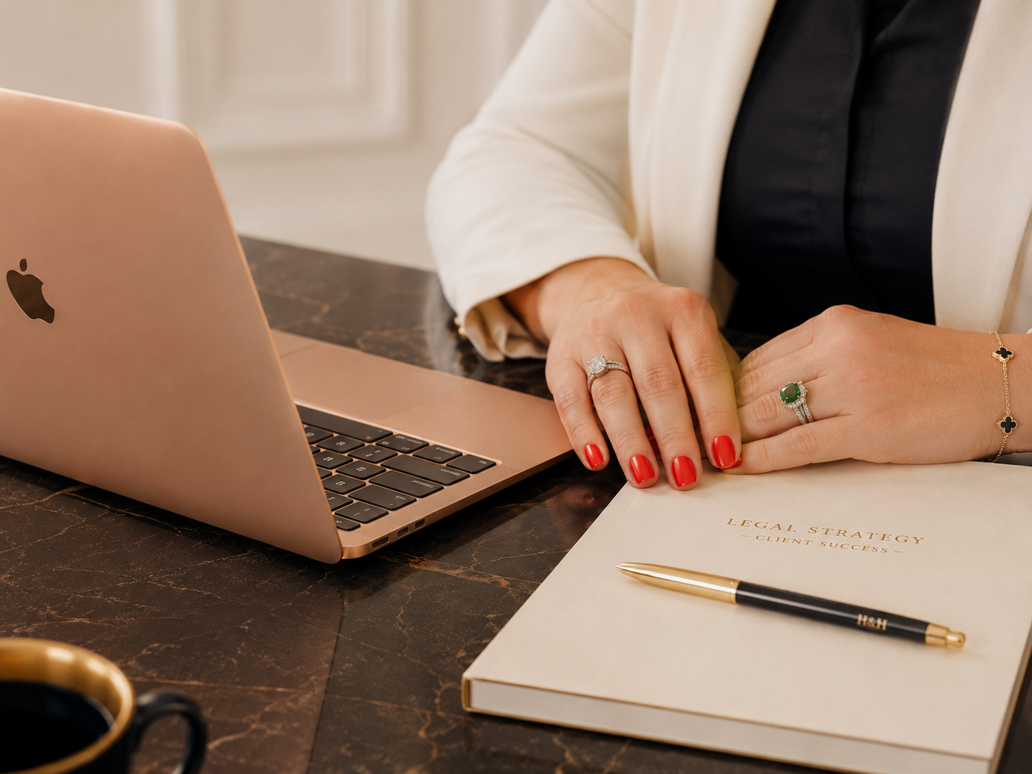 A close-up lifestyle shot of a woman's hands resting on a notebook with a pen, next to an open rose gold laptop. She is wearing a white blazer over a black top, with red nail polish and rings on her fingers.